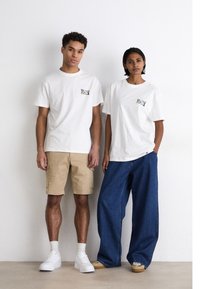 Dos modelos están de pie uno al lado del otro, vistiendo camisetas de algodón blancas con logotipos coloridos. Uno lleva un par de pantalones cortos caqui, el otro unos vaqueros anchos azules; ambos llevan zapatillas deportivas.