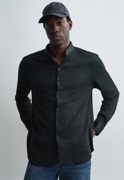Chemise noire à manches longues avec col mandarin, rayures verticales subtiles et boutonnage à l'avant. Associée à une casquette noire et un jean bleu.