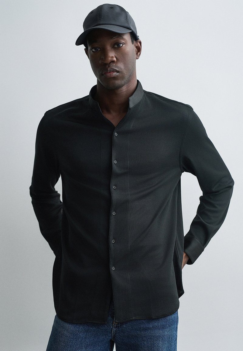 Mustassa pitkähihaisessa paidassa on mandariinikaulus, hienovaraiset pystyrivit ja napit edessä. Yhdistettynä mustaan lippikseen ja sinisiin farkkuihin.