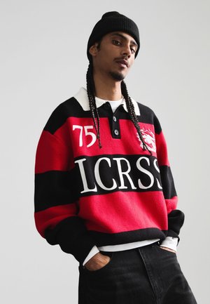 Fiatal férfi hosszú fonatokkal, fekete sapkában, piros-fekete-fehér csíkos pulóverben, amelyen a "75 LCRSS" felirat olvasható, kezét fekete nadrág zsebében tartja.