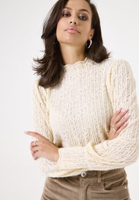 Crème gebreide trui met een textuurpatroon, ronde halslijn en geribbelde mouwen, gecombineerd met een paar bruine corduroy broek met hoge taille.