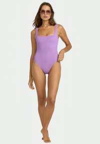 Maillot de bain une pièce violet à côtes avec un encolure carrée, des bretelles épaisses et une coupe mi-haute. Le modèle porte des lunettes de soleil et un collier en or.