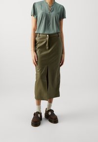 Hellgrün kurzärmlige Bluse mit rundem Ausschnitt, kombiniert mit einem hochgeschnittenen olivgrünen Lederrock mit Schlitz vorne und Knopf. Braune Schuhe.