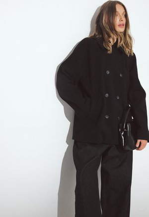 Zwarte double-breasted jas met grote knopen, voorzien van een gegroefde kraag en zijzakken, gecombineerd met donkere wijde pantalons en een glanzende handtas.