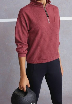 Femme portant un sweat-shirt à col montant zippé texturé bordeaux et un legging noir, tenant un ballon médicinal noir devant un fond neutre.