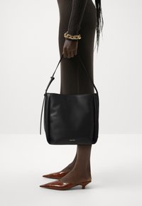Borsa in pelle nera con una sola tracolla, texture liscia e un logo prominente nella parte inferiore. Indossata con décolleté marroni a punta.