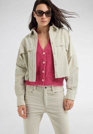 Donna con grandi occhiali da sole, giacca e pantaloni in velluto a coste color crema, con un gilet rosa peloso abbottonato, in piedi davanti a uno sfondo chiaro uniforme.