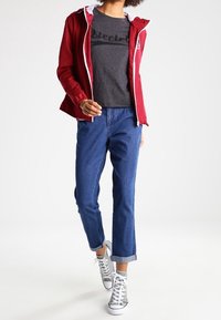 Červená lehká bunda s bílým lemováním, šedé tričko s černým textem, modré zkrácené džíny a stříbrné vysoké tenisky.