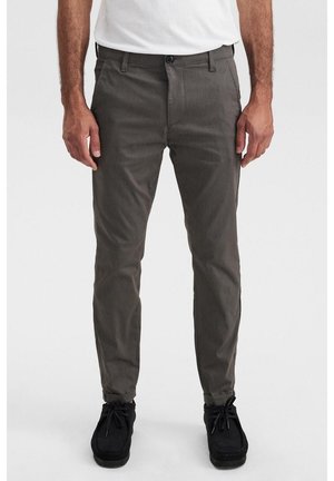 Uomo che indossa pantaloni slim fit grigio scuro, scarpe nere con lacci e camicia bianca, in piedi davanti a uno sfondo chiaro e uniforme.