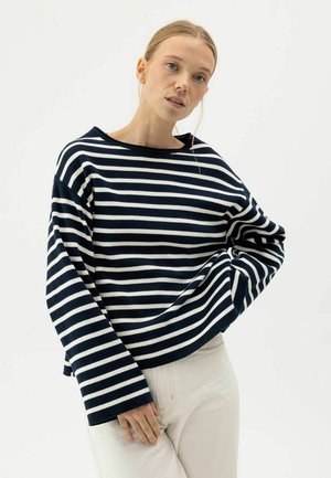 Sieviete ar blondiem matiem, tērpusies melnā un baltā horizontālu svītru garroku topā un gaišu biksēs, pozējot pret baltu fonu.