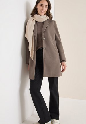 Brauner Wollmantel mit zwei Knöpfen, beiger Schal, brauner Pullover und schwarze Jeans. Das Model steht vor einer hellen Wand.