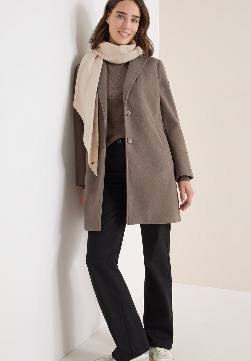 Manteau en laine marron avec deux boutons, écharpe beige, pull brun et jeans noirs. Le modèle se tient contre un mur de couleur claire.