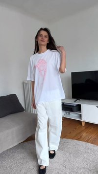 Jauna sieviete baltās biksēs un pārizmērā baltā T-kreklā ar rozā grafiku stāv telpās netālu no dīvāna un televizora, pieskaroties matiem.