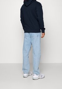 Sudadera de marina con un logo rojo, combinada con jeans de azul claro y zapatillas blancas. Diseño con capucha, material de algodón, corte relajado.