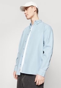 Chemise à manches longues bleu clair avec col, dotée d'une couche intérieure blanche et d'un logo de marque discret sur la poitrine. Texture de tissu douce.