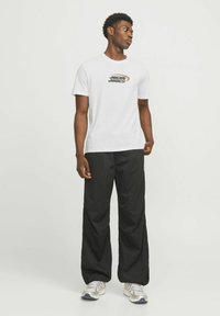 Witte T-shirt met een zwart en oranje grafisch logo, gecombineerd met losse zwarte broeken en grijze sportschoenen.
