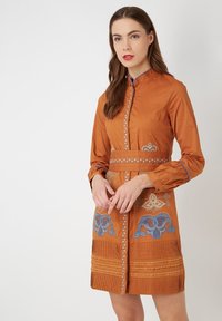 Vestido marrón de manga larga con cuello alto, presenta patrones bordados en azul y oro, una cintura entallada y acentos plisados en el dobladillo.