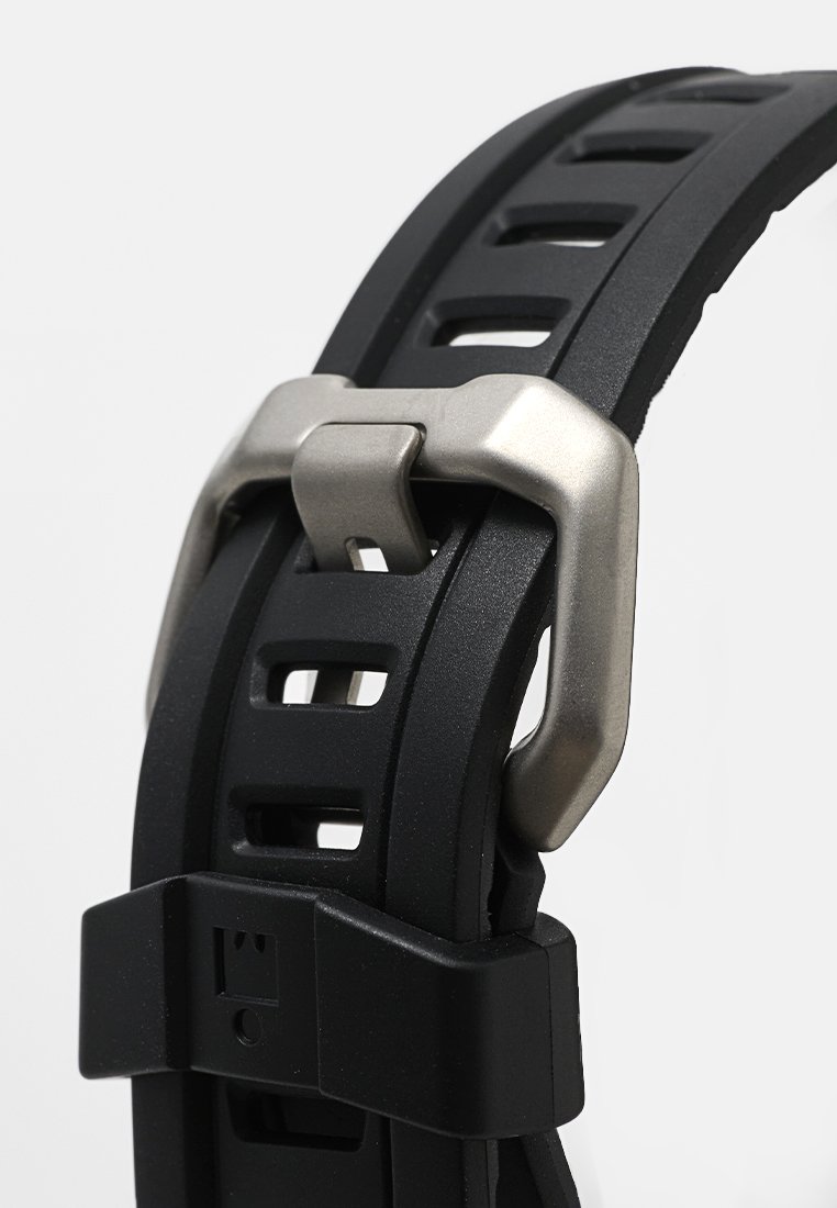 Correa de reloj de caucho negro con ranuras cortadas, que presenta un hebilla de metal plateado y un clasp de plástico. Superficie texturizada para mayor comodidad.