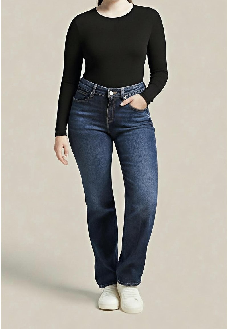 Personne portant un jean taille haute bleu foncé, un haut noir à manches longues, des baskets blanches, debout avec une main dans la poche sur un fond uni.