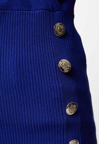 Tessuto a maglia blu rigato con bottoni rotondi dorati che presentano un emblema decorativo lungo un lato. Texture liscia con linee verticali.