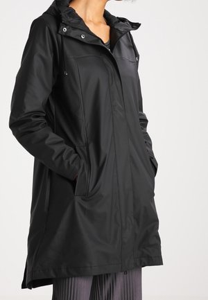 Manteau noir imperméable à capuche avec poches avant, porté sur un haut rayé et un pantalon plissé, présenté sur une personne debout de la nuque aux genoux.