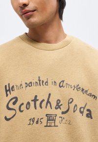 Άνδρας φοράει μπεζ φούτερ με μαύρο κείμενο «Handpainted in Amsterdam Scotch & Soda 1985» και μικρό γραφικό στο στήθος σε λευκό φόντο.