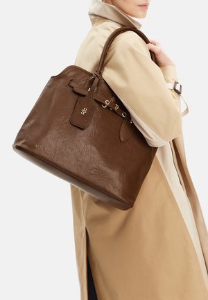 Mujer con gabardina beige llevando un gran bolso tote de cuero marrón con herrajes dorados en el hombro.