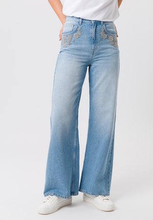 Persoon draagt lichtblauwe wijde jeans met decoratieve kralen op de voorzakken en witte sneakers, staand tegen een effen achtergrond.