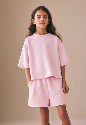 Junges Mädchen mit langen Haaren, das ein lockeres rosa Kurzarmoberteil und passende Shorts trägt und vor einem einfarbigen beigen Hintergrund steht.