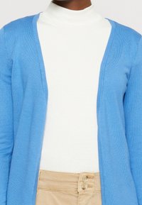 Ljusblå cardigan med en mjuk textur, öppen framsida och långa ärmar, kombinerad med en vit polotröja. Sett med beige byxor.