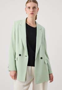 Blazer de corte cruzado en verde menta con botones negros y bolsillos, combinado con una blusa negra y pantalones color crema. Textura suave.