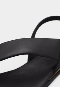 Μαύρο δερμάτινο σανδάλι με φαρδιά μπροστινή λουρίδα και στρογγυλεμένη πίσω λουρίδα πάνω σε επίπεδη σόλα σε λευκό φόντο.