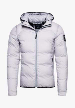Personne portant une doudoune gris clair, un pantalon cargo noir avec poches à fermeture éclair, un t-shirt noir et des baskets blanches, debout contre un mur blanc.