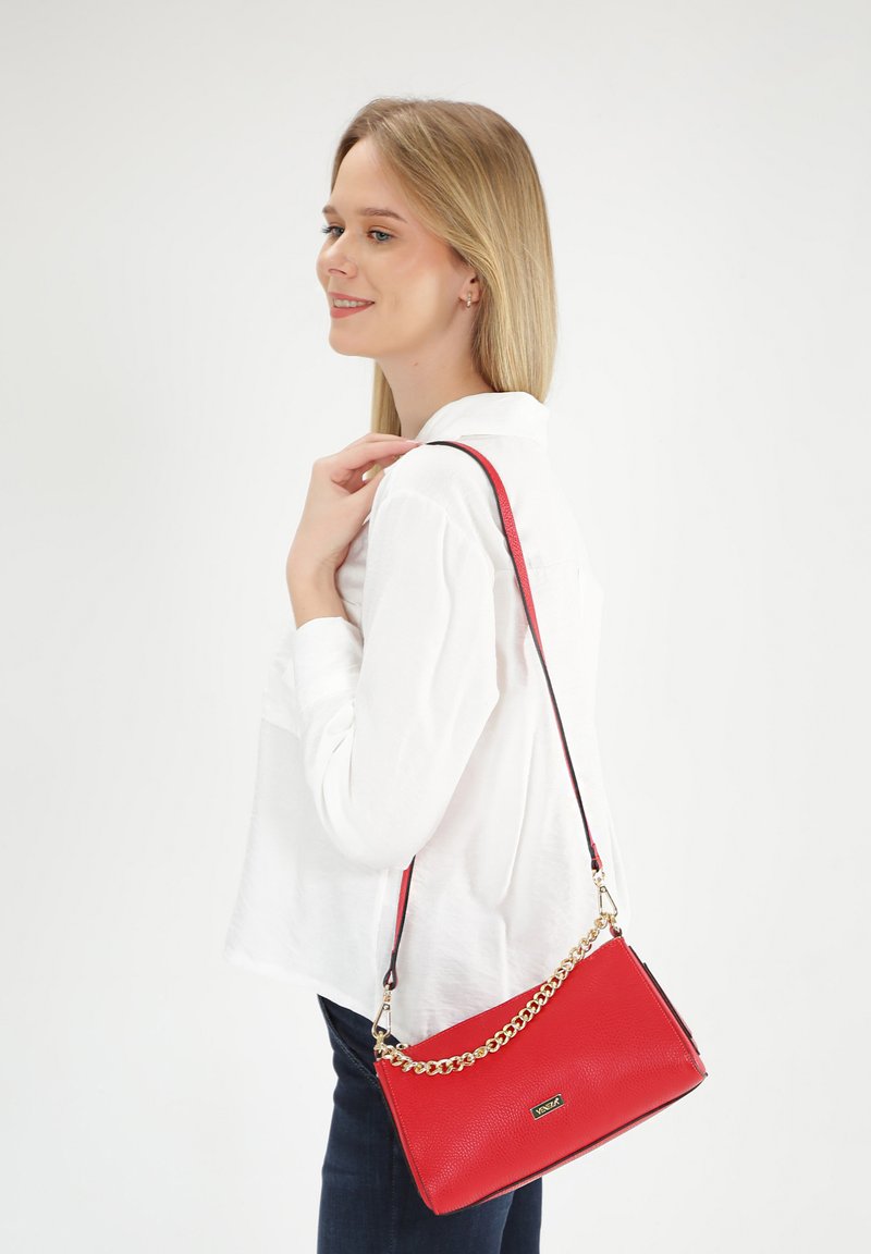Rode leren crossbody tas met een gestructureerde oppervlakte, gouden schouderband en zwarte accenten, vastgehouden door een persoon in een wit shirt.