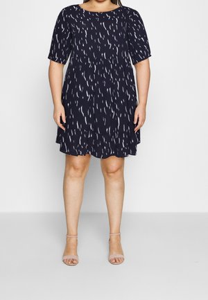 Žena v námořnicky modrých šatech po kolena s krátkými rukávy a bílým abstraktním vzorem, doplněná tělovými sandály s páskem kolem kotníku.