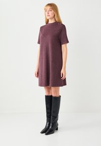 Krótka bordowa sukienka z krótkimi rękawami, wysokim dekoltem i rozcięciami po bokach. Z dwoma guzikami na ramieniu. Noszona z czarnymi kozakami do kolan.