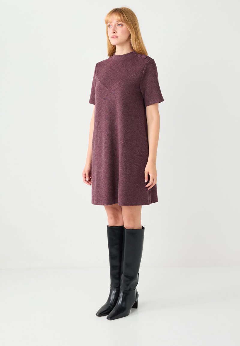 Krótka bordowa sukienka z krótkimi rękawami, wysokim dekoltem i rozcięciami po bokach. Z dwoma guzikami na ramieniu. Noszona z czarnymi kozakami do kolan.