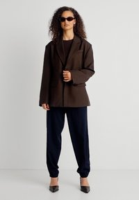 Γυναίκα με σγουρά μαλλιά που φοράει μαύρα γυαλιά ηλίου, υπερμεγέθη σκούρο καφέ μπλέιζερ, μαύρο παντελόνι και μαύρες μυτερές γόβες, στέκεται μπροστά σε λευκό φόντο.