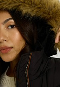 Jeune femme portant une veste noire avec une grande capuche bordée de fourrure brune, tenant la capuche près de son visage.