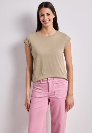 Lächelnde Frau steht frontal, trägt ein beiges Top mit kurzen Ärmeln und hoch taillierte pinke Jeans vor einem schlichten hellen Hintergrund.
