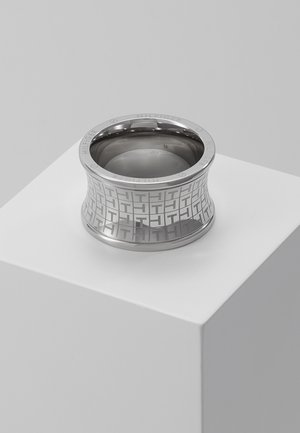 Brede zilveren metalen ring met gegraveerd herhalend patroon van "TH" en inscriptie "Hilfiger", geplaatst op een wit voetstuk tegen een grijze achtergrond.