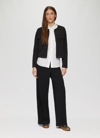 Veste noire courte avec deux poches avant, portée sur une chemise blanche et un pantalon large noir. Chaussures marron avec une finition texturée.