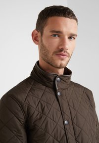 Casaco acolchoado castanho com gola alta, apresentando detalhes cinzentos e botões de pressão, exibindo um padrão de diamante texturizado.