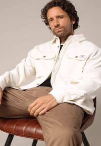 Veste en velours côtelé blanc avec poches poitrine, accents de boutons en argent et texture côtelée, portée avec un pantalon marron clair sur une chaise en cuir marron.