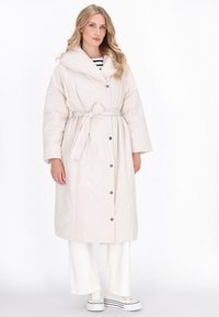 Palton pufos lung, de culoare roz deschis, cu un guler larg, talie evidențiată de un cordon și nasturi cu capsa. Purtați peste pantaloni albi cu croială largă și teniși albi.