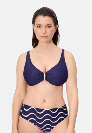 Mujer con top de bikini azul oscuro con cierre frontal y braguitas de bikini azul marino con patrón de olas blancas, de pie contra un fondo liso.