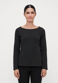 Woman with dark hair tied back wearing a black long-sleeve top and black pants, standing against a plain white background.