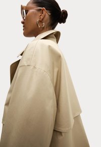 Beige oversized trenchcoat met een kraag, gladde textuur en verlaagde schouders. Vergezeld van duidelijke oversized brildragers en creolen.