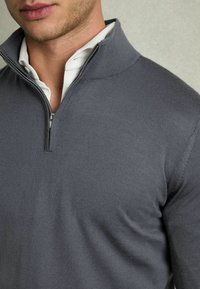 Grauer Pullover mit einem Viertelreißverschluss-Kragen und geripptem Struktur. Verfügt über einen glatten Stoff und einen darunterliegenden weißen Kragen.