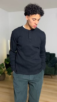 Fekete, bordás mintájú hosszú ujjú ing gombos gallérral. Laza, türkizkék kord nadrággal viselve. A háttérben zöld kanapé és növény látható.